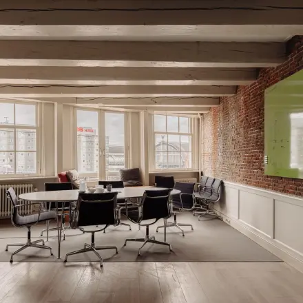 Charming office space at Prins Hendrikkade 14 in Amsterdam Center with exposed brick walls, a large table, modern chairs, and a large window with city views. Ideal for office space rental.