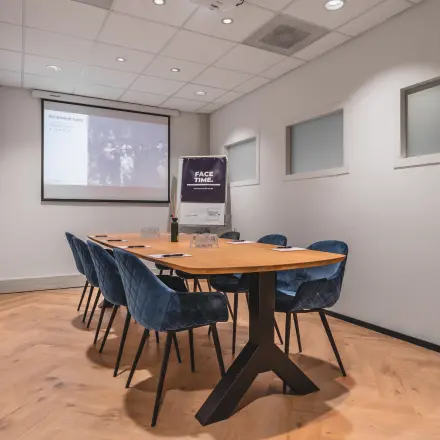 A conference room featuring a sleek wooden table with blue chairs, a large screen, and projector setup, perfect for office space rental in Amsterdam Duivendrecht.