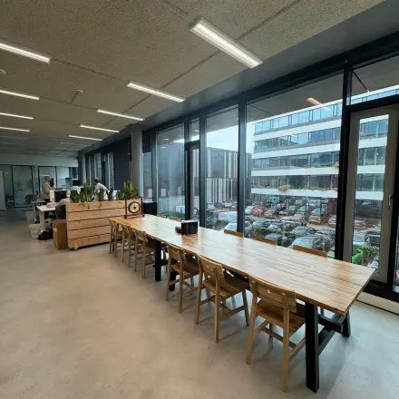 Open office space at Moermanskkade 303 in Amsterdam Houthavens, featuring a long wooden table with chairs and large windows. A person works at a desk in the background.