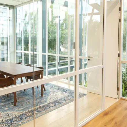 Bright office space with a large wooden table and chairs surrounded by glass walls offering views of greenery, ideal for office space rental.