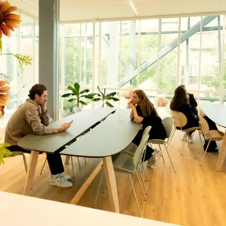 Office space rental in Amsterdam Riekerpolder, featuring spacious tables and large windows. People are engaged in conversation, adding a collaborative vibe.