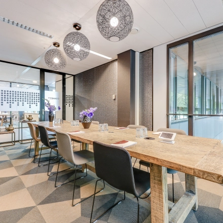 Bright office space in Amsterdam Riekerpolder with a wooden table, modern chairs, and glass partitions, ideal for rent office space. The setting is modern with hanging lights and natural lighting.
