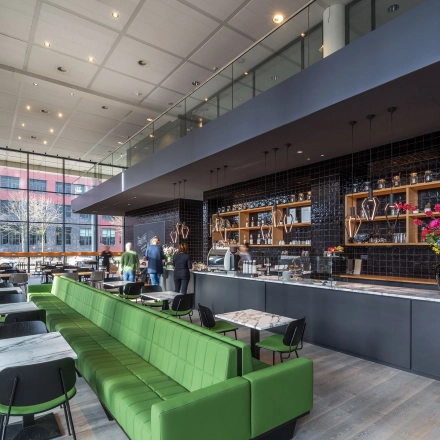 Modern office at Kingsfordweg 43, Amsterdam Sloterdijk, featuring an open lounge area with green seating and tables. People are engaged in conversation near the counter, enhancing the vibrant office space rental.