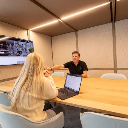 Two people engage in a meeting in a modern, well-lit office space with a large wooden table and digital screen, suitable for office space rental in Amsterdam South East.