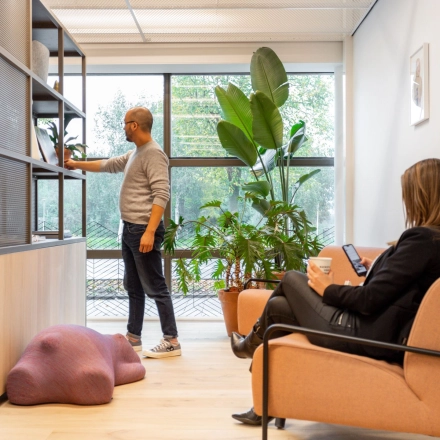 A modern office at Haarlemmerweg 331, Amsterdam West, with a person browsing shelves and another seated with a tablet. Ideal for office space rental.