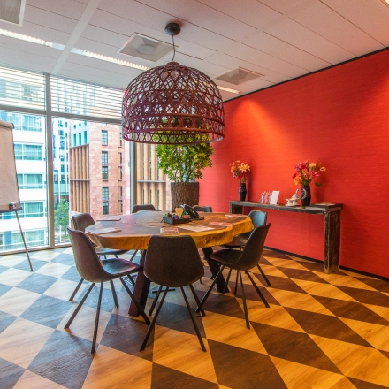 Bright conference room in Amsterdam Zuidas, featuring a large round table with chairs, vibrant red walls, and a large window overlooking a cityscape. Ideal for office space rental.