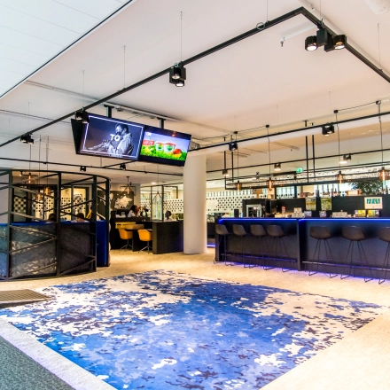 Bright, furnished office space available for rent at Gustav Mahlerplein in Amsterdam Zuidas, featuring a stylish open floorplan with a vibrant blue carpet and modern seating area.