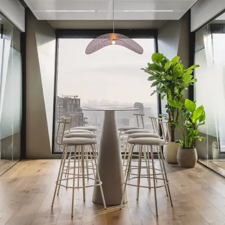 A modern office space with tall windows in Amsterdam Zuidas, featuring high stools around a central table, ideal for collaborative meetings. Rent office space with stunning city views.