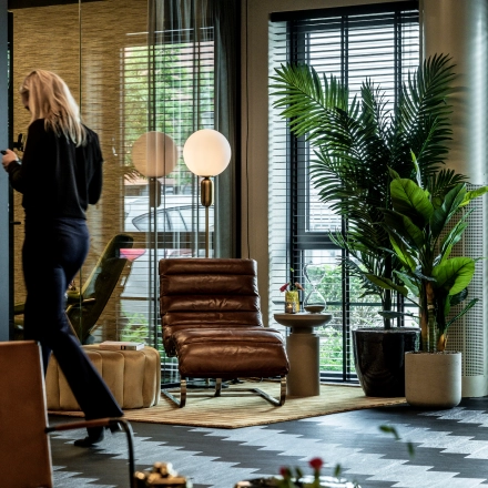 Modern furnished office space in Apeldoorn with stylish seating and plants. A person is walking while using a smartphone, suggesting business activity.