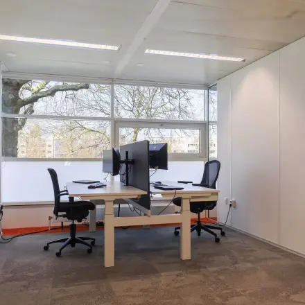 Bright office space for rent in Apeldoorn, featuring two desks with chairs and screens, large windows offering natural light.