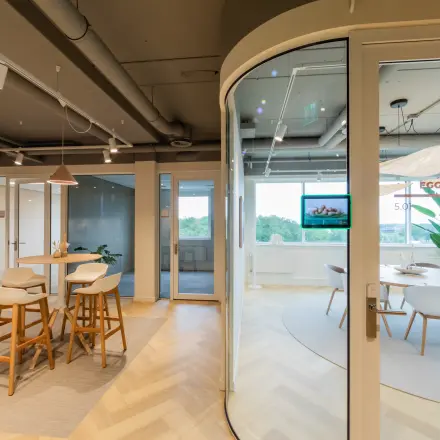 Modern office space for rent featuring glass-walled meeting room, bar stools at a high table, and large windows providing natural light.