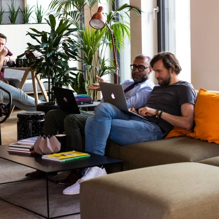 Business professionals collaborating on laptops in a modern office lounge with stylish furniture and indoor plants at Warschauer Platz 11-13, ideal for office space rental in Berlin Friedrichshain.