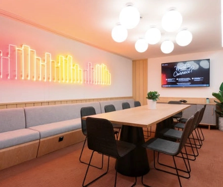 Conference room at Warschauer Platz 11-13, Berlin Friedrichshain, featuring a long wooden table surrounded by chairs, neon wall art, and a large screen. Ideal office space rental for meetings.