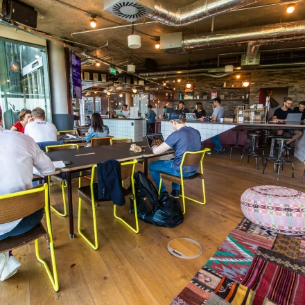 Modern office space rental at Warschauer Platz 11-13, Berlin Friedrichshain, showcasing individuals collaborating over a large table with vibrant decor and industrial lighting.