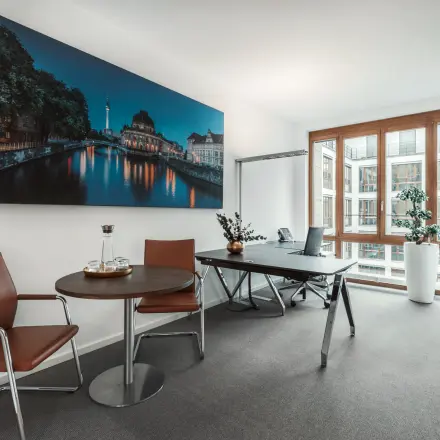 Modern Berlin Mitte office space rental at Pariser Platz 6a, featuring a meeting area with chairs, table, and work desk near large windows.
