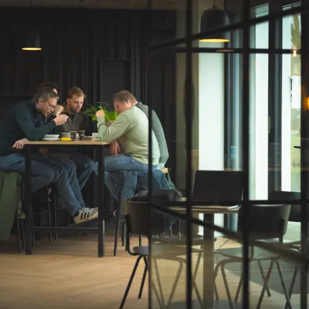 Three people sit at a tall table in a modern office space at Rietbaan 2-12, enjoying a meal while discussing business. The room is stylishly furnished with large windows.