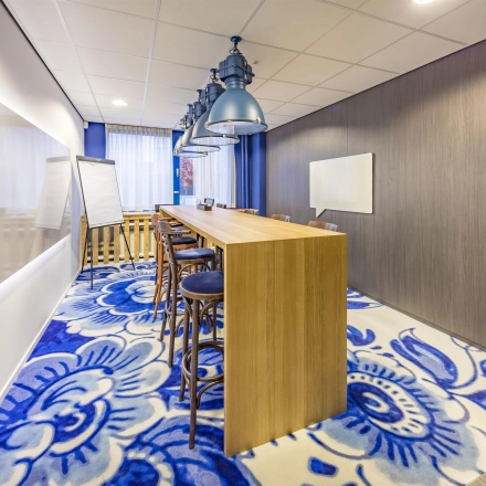 Brightly lit office space with a long wooden table and chairs on a floral carpet at Amerikastraat 3, Den Bosch, ideal for office space rental.