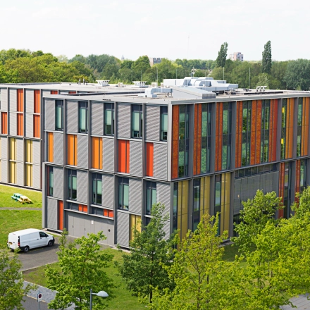 Exterior view of a modern, colorful office building at High Tech Campus 10, Eindhoven, surrounded by greenery with parked vehicles nearby. An ideal location for office space rental.