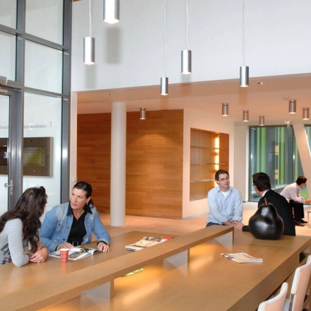 Modern office lounge at High Tech Campus 9, Eindhoven with natural light, offering comfortable seating and tables ideal for business meetings. People are engaged in discussions, perfect for office space rental.