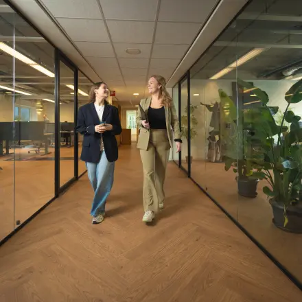 Two people walking and conversing in a modern office to rent at Brouwerijstraat 1, featuring glass walls and wooden flooring with office plants.