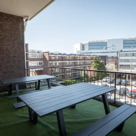 Outdoor office space rental at Kuipersdijk 2-4 in Enschede, featuring two black picnic tables on artificial grass, overlooking a parking area and adjacent buildings.