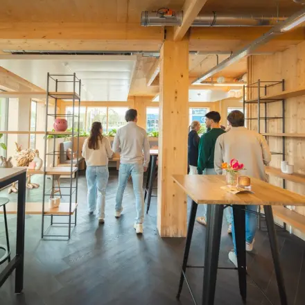 Modern office space at Moutlaan 1-23, Enschede, featuring stylish wooden interiors with shelves and high chairs, ideal for office space rental. A group of people discusses business strategies.