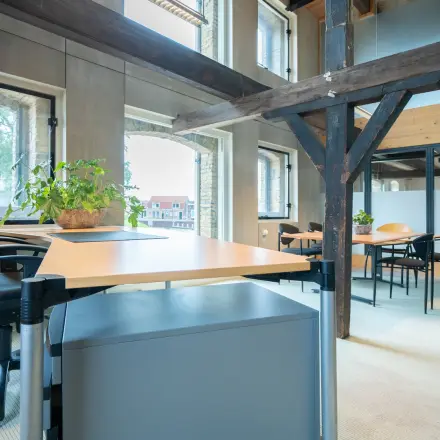 Office space rental at Prins Hendrikkade 3, Franeker, featuring multiple workspaces with desks and chairs, natural light from large windows, and industrial beams.