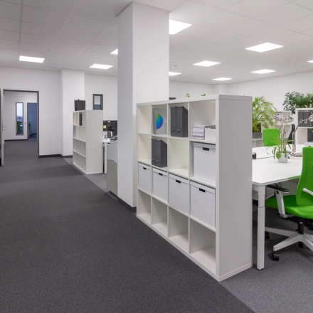 Modern office space in Frankfurt West featuring green chairs, ample shelving, and large desks for workspace rental. Bright and inviting atmosphere with overhead lighting and large windows.