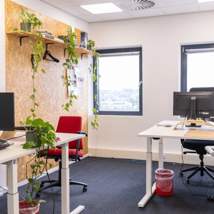 Bright, furnished office space in Gouda with desks and plants, ideal for office space rental. Neutral decor with large windows providing natural light and a view. Two red chairs add a pop of color.