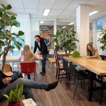 A furnished open-plan office space at Waarderweg 19, Haarlem Waarderpolder, features people working at tables surrounded by greenery. Ideal for those seeking office space rental.