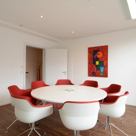 Bright office space with red chairs around a white table, ideal for meetings. Modern decor includes abstract art on the wall. Perfect for rent office space inquiries.