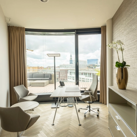 Bright office space rental in Hamburg Neustadt with modern furnishings, featuring a desk, chairs, and a view of the cityscape through large windows.