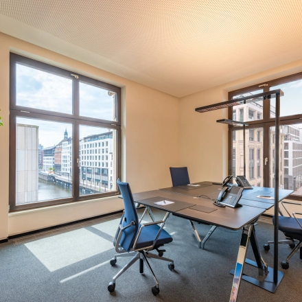 A bright, furnished office to rent at Alter Wall 32, Hamburg Old Town, featuring large windows, a meeting table, and chairs overlooking the cityscape.