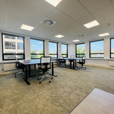 Bright office space in Hilversum featuring multiple black desks and mesh chairs by large windows with a view, available for rent.