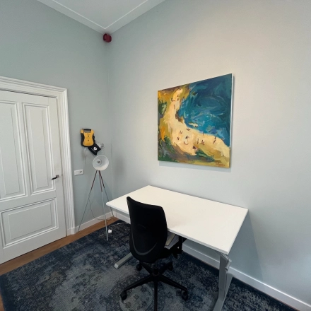 Small, furnished office space with a white desk, black chair, and colorful artwork on the wall, available for office space rental in Hilversum.