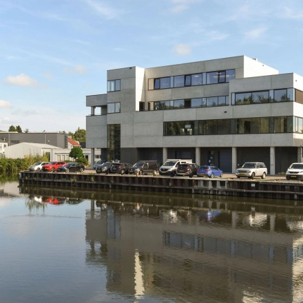 Exterior view of an office building at Zuiderloswal 55 in Hilversum, reflecting in a nearby canal. Multiple cars are parked outside. Rent office space in a scenic location.