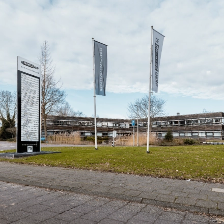 Exterior view of an office building at De Vos van Steenwijklaan 75, Hoogeveen, surrounded by greenery with signage and flags, offering office space rental options.