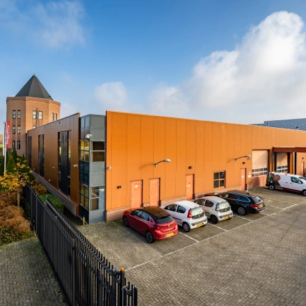 Exterior view of a modern office building at Hoofdveste 2, Houten, featuring an orange facade with a parking area in the foreground. Ideal for office space rental, this location offers convenience and accessibility.