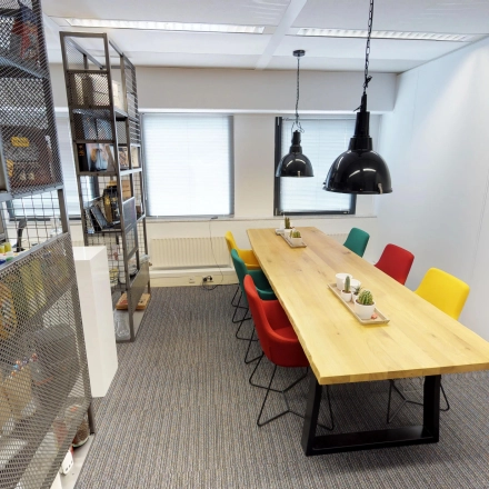 Bright office space at Meidoornkade 22, Houten, featuring a long wooden table with colorful chairs, ideal for meetings. Shelves line one wall, adding functional storage.