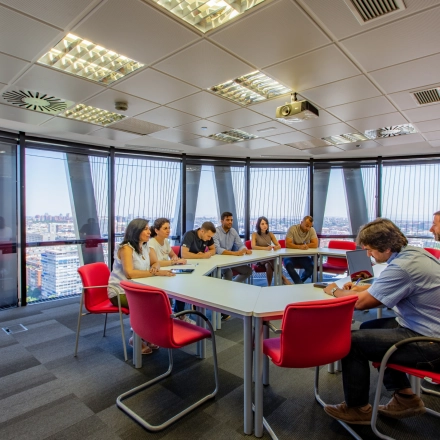 A conference room in Paseo de la Castellana 95, Madrid Azca, with people engaged in a meeting around a large table, featuring large windows and red chairs, available for office space rental.