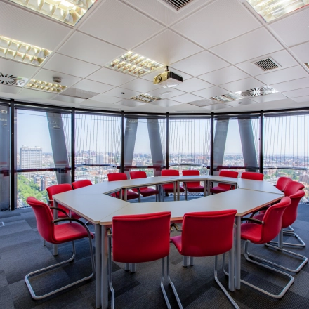 Bright office space with a panoramic city view, featuring a round conference table surrounded by red chairs, ideal for meetings or presentations.