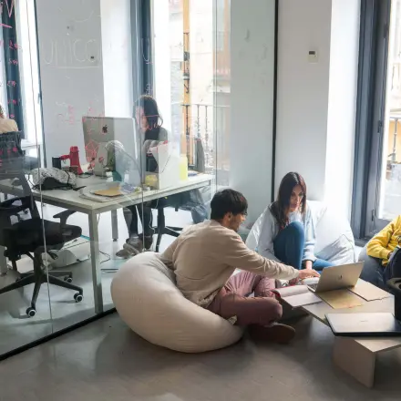 Office space to rent in Madrid Center with modern furnishings, featuring people collaborating around a laptop and others working at desks in a bright setting.
