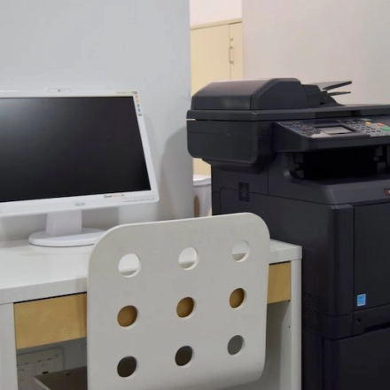 Compact office equipment setup with a desk, chair, monitor, and multifunction printer, ideal for those seeking office space rental in Madrid Center.
