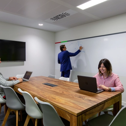 Professionals engage in business activities in a furnished office space with a conference table and a wall-mounted screen, ideal for office space rental in calle de O'Donnell, Madrid Retiro.