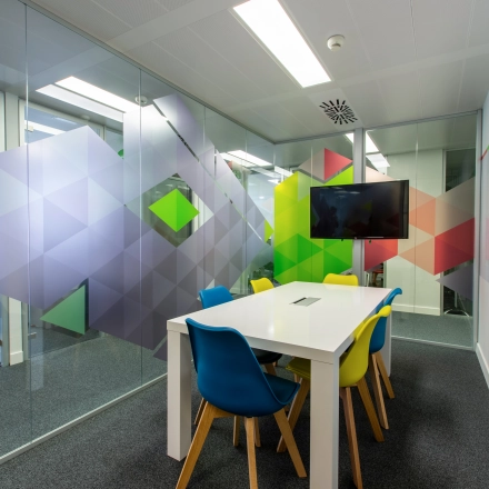 Modern office space rental at Calle de O'Donnell, Madrid Retiro. Features colorful chairs surrounding a white table in a meeting room with vibrant glass wall designs.