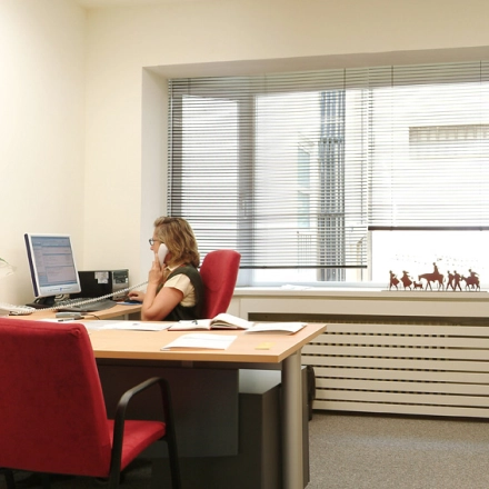 A furnished office space for rent on Avenida de Brasil in Madrid Tetuán, featuring a desk, red chairs, and a person working on a computer by the window.