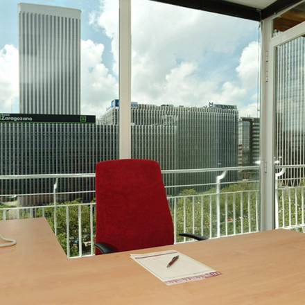 An office to rent in Madrid Tetuán with a desk, chair, and telephone, offering a view of city skyscrapers through large windows.