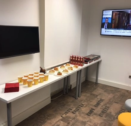 Cozy office lounge with a long table set for snacks, beverages, and a wall-mounted TV. Ideal for office space rental in Calle Capitan Haya 60, Madrid Tetuán.