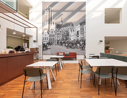 Bright office space in Mechelen Centrum with modern furniture and a large mural, ideal for office space rental. Tables and chairs arranged for collaborative work.