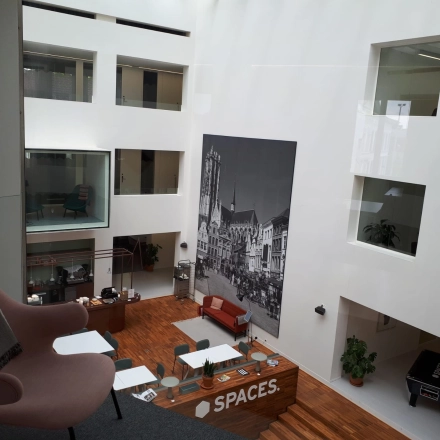 A modern office space rental in Mechelen Centrum featuring a bright, open atrium with a seating area and large artwork on the wall. Ideal office to rent for meetings or collaboration.